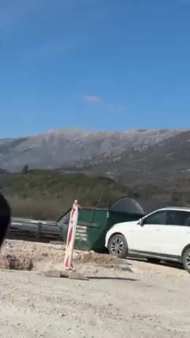 The video shows a scene of construction work taking place against the backdrop of a mountainous landscape under a clear blue sky. The camera is positioned inside a vehicle, with the view captured through the passenger side window.

In the foreground, there are construction materials, including red and white striped barriers, concrete blocks, and a green dumpster. A white car is partially visible next to the dumpster.  

As the vehicle moves forward, a concrete mixer truck comes into view, parked near a newly constructed concrete structure. A construction worker wearing a yellow vest is standing near the truck. 

In the background, forested hills transition into larger mountains with some snow covering their peaks. The overall scene suggests construction work is underway in a scenic, rural area.