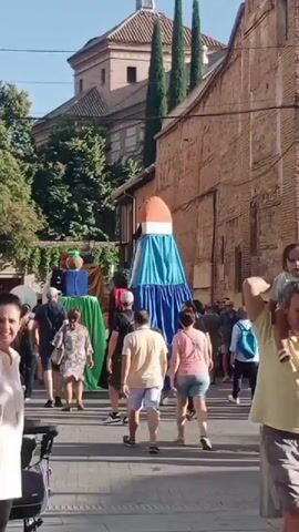 The video shows a street scene during what appears to be a festival or parade. In the center of the frame, a large, colorful figure resembling a rocket or a giant puppet is visible, dressed in blue and white fabric with an orange top. The figure is standing upright in a street lined with brick buildings and trees. A crowd of people, including adults and children, are walking around and observing the figure. Some individuals are pushing strollers. The overall atmosphere seems festive and crowded. The lighting suggests it is daytime, likely late afternoon or early evening, given the warm light and shadows. A woman with dark hair and a light-colored shirt is in the foreground, partially obscuring the view of the central figures. Buildings in the background feature architectural details, including a domed roof on one structure. The ground is paved with cobblestones or similar material.