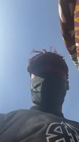The video captures a close-up, low-angle selfie of an individual outdoors under a bright, clear blue sky. The person, who appears male, is wearing a black face mask, a distressed brown cap with "NEW YORK" text, and a black long-sleeved shirt featuring a white graphic on the lower right.

Throughout the initial frames, only their eyes are visible above the mask. Around the 0:02 mark, they pull the mask down with their right hand, revealing a broad, friendly smile. Their left hand holds a black cable, possibly from headphones or earbuds.

The background shows distant trees, including a prominent tall, slender one, and several small, simple structures on reddish-brown earth, suggesting a rural or semi-rural setting. Strong wind noise is distinctly audible, indicating breezy conditions. The bright sunlight suggests it is daytime. The general environment is consistent with the provided context of Mode, Nigeria.