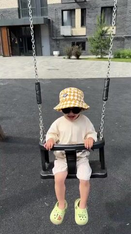 A child wearing a checkered bucket hat and sunglasses is sitting on a swing. The child is wearing a light-colored t-shirt and striped shorts. The child is smiling and appears to be enjoying the swing. The swing is in a playground with a building in the background. The ground is covered in a dark, soft material. The child swings forward and backward. The child is holding onto the sides of the swing. The child's feet are dangling in front of them. The child's mouth is open as if speaking or laughing.