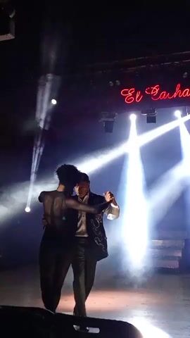 A couple dances on a dimly lit stage. They are illuminated by bright white spotlights from above, which create beams of light cutting through the smoky atmosphere. The woman, on the left, wears a dark, sheer, form-fitting outfit that reveals her silhouette. Her arms are around the man's neck, and she leans into him. The man, on the right, is dressed in a dark suit with a patterned jacket and a light-colored shirt. He holds the woman's waist with his left arm and her right hand with his left hand. They are facing each other and appear to be engaged in a close embrace, possibly dancing a tango or similar partner dance. Above and behind them, a neon sign in cursive red letters spells out "El Bachata". The stage floor is dark and reflective, catching the light. The background is dark, with some stage lighting rigs visible at the top. The bottom of the frame shows a dark foreground, possibly the edge of the audience or stage barrier.