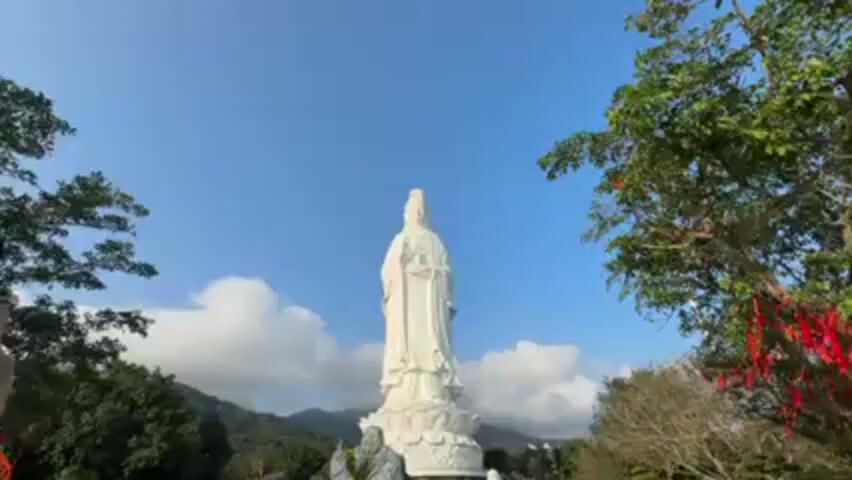 The video shows a full shot of a large, white Buddha statue in a garden. The statue is tall and imposing, set against a clear blue sky with some scattered clouds. The surrounding garden is filled with trees, some of which are adorned with red ribbons. In the background, there are mountains. The camera slowly pans to the right. There are several people in the scene, but they are out of focus and partially obscured by the statue.