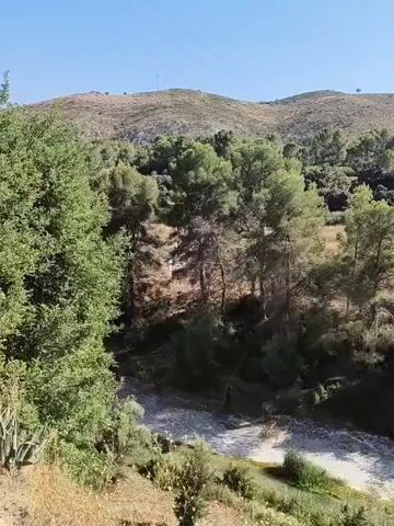 A drone flies over a dry riverbed lined with trees and brush. The riverbed is a pale, rocky expanse. The surrounding terrain is hilly and covered in vegetation, with more trees in the middle ground and a distant view of a mountain range under a clear blue sky.