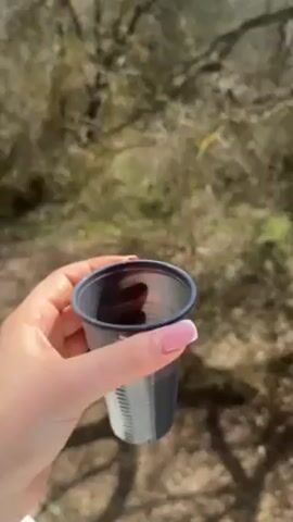 Here is a description of the video:

A person holds a small black and white disposable cup containing a liquid that appears to be coffee. The cup is held at eye level, and the background reveals a natural outdoor setting with trees, grass, and a hint of blue sky. The trees are bare, suggesting it might be late fall or early spring. The person's hand is visible, showing manicured nails with a pink and white design. The camera angle emphasizes the cup and the surrounding nature.
