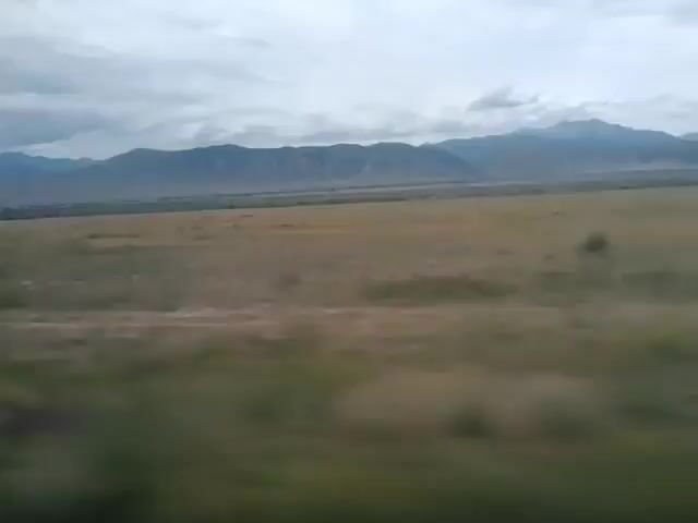 A time-lapse video shows a vast, open plain with rolling hills stretching towards a mountain range in the distance under a cloudy sky. The camera is moving, giving the impression of travel. The terrain is mostly covered in dry grass and scattered patches of greenery. The mountains are hazy and appear to be quite far away. The clouds are varied, with some appearing darker and more ominous than others. There are no people or man-made structures immediately visible in the foreground, though there are faint signs of development in the distance near the base of the mountains. The overall mood is serene and expansive.