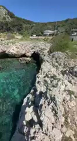 The video presents a panoramic view of a beautiful, rocky coastline under a clear blue sky. The scene is dominated by the strikingly clear, turquoise-to-emerald green water of the sea, revealing rocks and the seabed beneath.

The camera begins by showing the water close to the rocky shore, then slowly pans to the right, revealing more of the vast, calm sea stretching towards the horizon. In the distance, several dark, elongated shapes, possibly small boats or rocks, are visible on the water's surface. The coastline consists of rugged, light-colored rock formations that jut into the sea, forming small coves and inlets.

As the pan continues, a distant landmass becomes visible on the right, featuring low hills covered with sparse green vegetation. A lighter-colored structure, possibly a building or ruins, can be seen on top of one of these distant hills. The bright sunlight suggests it is daytime, likely mid-morning or afternoon, with excellent weather conditions. No people or explicit human activity are visible, emphasizing the natural beauty and tranquility of the coastal landscape. The overall impression is one of serene, unspoiled natural beauty in a sunny, coastal environment, typical of regions like Nardò, Italy.