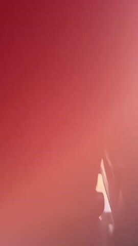 The video opens with a blurry, reddish-pink frame, transitioning to the interior of a car. A person's hand is visible inside the vehicle, with the back of a car seat and a glimpse of another person's blonde hair in the background. The scene then becomes very bright and blurry inside the car before shifting to an exterior view from the car window. The car is driving along a road, next to a silver guardrail. Beyond the guardrail, lush green hills and trees are visible under a clear, bright blue sky, indicating a sunny day. No text is visible. The scene suggests driving through a green, natural landscape.