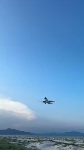 A commercial airplane flies low in the sky over a beach and ocean. The airplane has its landing gear down. The sky is clear and blue with a few clouds. The sun is low in the sky, casting a warm light. Palm trees and mountains are visible in the distance. There is a person standing on the beach taking a photo of the airplane.