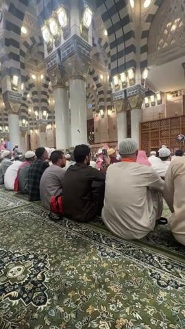 The video shows a wide shot of the interior of a mosque. Many people are seated on the floor, facing a raised platform. They are dressed in traditional Islamic attire. The mosque features elaborate architecture with striped columns and ornate ceilings. The lighting is warm and atmospheric. The camera pans slowly across the scene, capturing the serene atmosphere of the gathering.