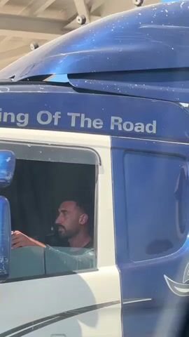 This is a close-up, medium-shot video of a large blue and white truck, likely a semi-trailer or transport vehicle. The truck is parked under an overhead structure, possibly a covered waiting area or a toll booth, given the presence of what appears to be a metal roof with support beams. Sunlight suggests it's daytime, and the clear conditions imply good weather.

The primary subject is a man with dark hair and a beard, who is seated in the driver's seat of the truck. He is wearing a green t-shirt and is looking towards the left side of the frame, his right hand resting on something inside the truck's cab. His expression is neutral.

A prominent detail on the truck's blue cab is the text "King Of The Road" in white lettering. The truck has large, reflective blue side mirrors, one of which is partially visible in the foreground. The overall impression is one of a routine moment, possibly a pause in a journey or waiting at a checkpoint.

The scene is likely taking place in Beit Horon, Palestinian Territories, as indicated by the provided location context. While there are no explicit visual cues in the video itself to confirm this precise location, the context informs the understanding of the setting as a transportation hub or route within that region. The video is short, with no significant action or change in scene.