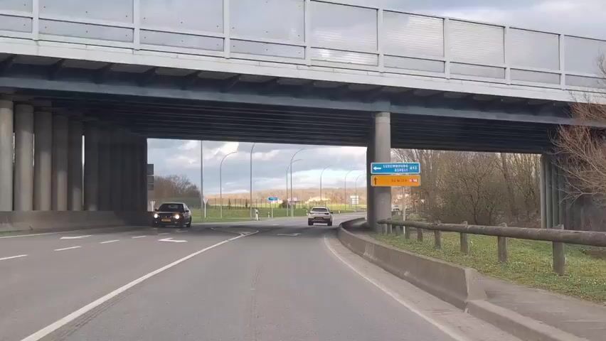A camera mounted on a vehicle records footage as it passes under an overpass. Two cars are visible ahead on the road. One car is turning right at a fork in the road, while the other car is proceeding straight. A road sign indicates that going left leads to Luxembourg A13 Aspe and going straight leads to Remich. The sky is overcast. Trees without leaves are visible in the background. The overpass is supported by many columns.