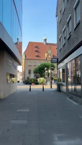 This is a vertical, eye-level shot of a pedestrian walkway leading towards a square. The day is bright and sunny, suggesting it is daytime. The architecture on either side of the walkway is a mix of modern and traditional. On the left, a modern building with a glass facade reflects the blue sky and surrounding buildings. On the right, a more traditional, older building has several windows and an awning extending over a shopfront.

In the background, a large, historic building with a red-tiled roof dominates the view. This building features a prominent clock face and appears to be a town hall or significant civic structure. The square in front of it has outdoor seating for a cafe or restaurant, with tables and chairs, and some potted plants. A lone person, a male wearing a light-colored t-shirt and dark shorts, is walking through the walkway towards the square. He is carrying a large bouquet of white flowers. Another person, a woman wearing a pink top and white shorts, is visible further in the square, walking away from the camera.

A sign attached to a lamppost reads "back WERK" in white lettering on a black background. The walkway is paved with gray stone tiles. Two black bollards are placed at intervals along the walkway. The overall mood is peaceful and pleasant, suggesting a calm, everyday scene in a European town.