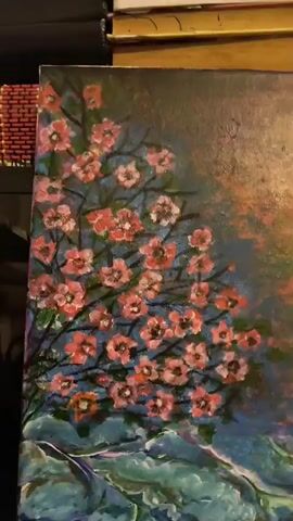 The video displays a close-up, slightly shaky shot of an acrylic painting. The painting features a cluster of pink and white cherry blossoms against a dark blue background, with a suggestion of water or rocks in shades of green and blue at the bottom. The upper right portion of the painting has a textured, blurry effect with hues of orange and red. The painting is positioned on a dark surface, possibly a keyboard, with a slight reflection visible on the right side. The lighting is dim, and the camera is not perfectly still.