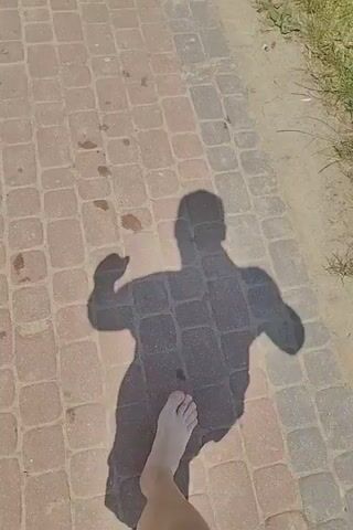 A shadow of a person walks across a paved walkway. The shadow is that of a muscular individual, with their arms bent and fists clenched. The walkway is made of light brown rectangular bricks. To the right of the walkway is a patch of dry grass and dirt. The sun is shining, casting a long shadow. A bare foot enters the frame from the bottom left corner and is visible in the subsequent frames, walking on the walkway. The shadow's movement suggests the person is walking forward.