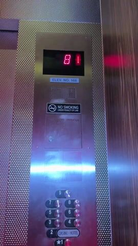The camera is static and pointed at an elevator panel. The panel is made of patterned metal and has a digital display showing the number 71 and a downward arrow. Below the display, there is a sign that reads "ELEV. NO. 166" and a "No Smoking" sign. Further down, there are rows of round buttons labeled 9, 7, 8, 5, 6, 3, 4, and 2, along with a button labeled "Casino Hotel" and a star symbol. The elevator is likely in a casino hotel, and is descending from the 71st floor. A vertical strip of dark wood is visible on the right side of the frame.