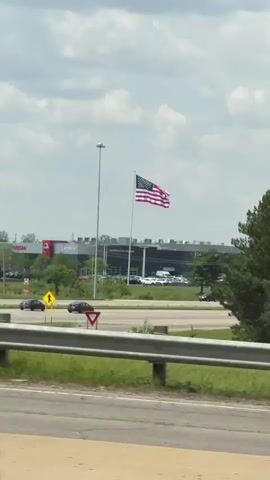 The video captures a daytime view from a moving vehicle, looking across a highway or main road towards a commercial area. In the foreground, a metal guardrail runs parallel to the road. Two road signs are visible on the left: a yellow diamond sign with a black arrow pointing left, and a red triangular sign with a black arrow pointing upwards and to the left.

Across the road, a large American flag flies high on a flagpole. Behind it, several commercial buildings are visible. One prominent building has a "NISSAN" sign, and another appears to be a "Chevrolet" dealership, though only partially visible. Several cars are parked in the dealership lots, and others are driving on the roads. A large light pole stands near the flag.

As the video progresses, a large, dense green tree in the middle ground partially obscures the buildings and the left side of the frame. The American flag remains visible above the trees. The sky is bright with scattered clouds. A soft "ooh" sound is heard at the beginning of the video.