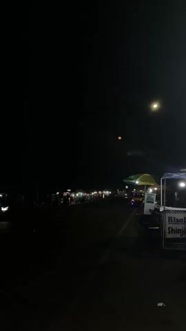 The video displays a nighttime street scene with multiple food stalls and people. The atmosphere appears lively, with the stalls illuminated by lights and several individuals visible, suggesting commercial activity and pedestrian traffic. The overall setting suggests an outdoor market or a street lined with vendors, likely a common area for social gatherings or evening dining.