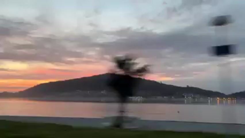 The video shows a scenic view of a body of water, possibly a bay or ocean, at sunrise or sunset.  The sky is filled with clouds painted in hues of orange, pink, and grey. A landmass, likely a mountain or large hill, is silhouetted against the vibrant sky in the background.  Lights from a settlement are visible along the shoreline of the landmass. The water is calm and reflects the colors of the sky.  The camera appears to be moving, possibly from inside a car or other vehicle.