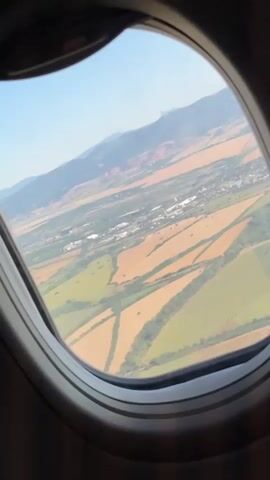 A view from an airplane window shows a landscape of agricultural fields. The fields are a mix of green and brown, with some areas appearing to be recently harvested. The fields are divided by dirt roads and lines of trees. In the distance, mountains can be seen. The sky is clear and blue. The video is a still image, with no visible movement or action.