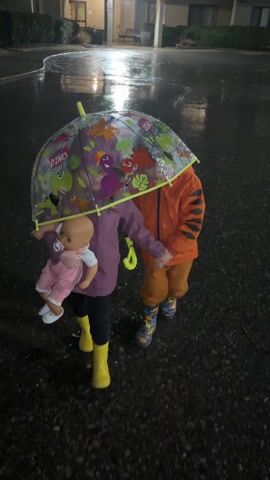 Two small children, one wearing a purple jacket and yellow boots and holding a doll, and the other wearing an orange jacket and multicolored boots, stand together under a clear, dinosaur-themed umbrella in the rain. They are holding hands. The ground is wet, and reflections from lights are visible in the puddles. The setting appears to be a parking lot or walkway adjacent to a building, possibly at night. Rain is falling heavily.