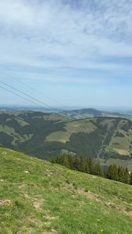 The video presents a scenic view of rolling hills and valleys under a partly cloudy sky. The foreground features a grassy slope dotted with yellow flowers and some pine trees. In the distance, a landscape of alternating green hills and forested areas stretches toward the horizon. Utility lines are visible in the upper left portion of the frame. As the video progresses, a small metal swing set comes into view on the hillside. The scene is shot from a slightly elevated position, presumably on a hilltop or ridge.