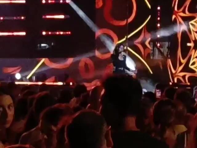 A live performance is taking place on a stage in front of a crowd. A female singer is on stage, wearing a black outfit, and she is holding a microphone. The stage has a large screen behind it with a colorful, abstract design and bright lights. Beams of light are shining across the stage and into the audience. The crowd is visible in the foreground, with many people watching the performance. The lighting is dim, creating a concert atmosphere. The video is shaky, suggesting it was filmed by someone in the audience.