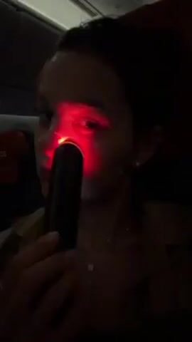 A person is sitting in what appears to be an airplane seat, using a handheld device that emits a red light on their face. The device is black and cylindrical, and the red light is concentrated on the person’s cheek. The person is wearing a light-colored shirt and has short dark hair. There is a face mask on the upper part of their nose. The lighting in the cabin is dim, and there is a headrest visible behind the person, indicating they are likely on a flight.