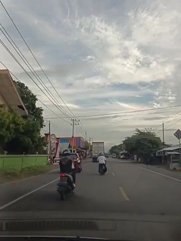 The video shows a street scene with several vehicles and buildings. The sky is cloudy, with wispy clouds. On the left side of the road, there are some trees and buildings. One building is red and blue, and another is a small shop. There are also utility poles with many wires. On the right side of the road, there are more buildings and trees.

Several vehicles are on the road. In the foreground, a car is driving, as indicated by the dashboard at the bottom of the frame. A motorcycle is in front of the car, with two people on it. Further down the road, there are more motorcycles and cars. A large truck with a blue tank is also visible. The road has yellow lines marking the lanes. The scene appears to be a busy street in a town or city, possibly in Southeast Asia due to the presence of many motorcycles and the style of the buildings. The time of day seems to be morning or late afternoon, given the lighting conditions.