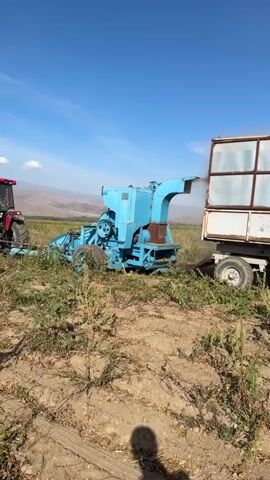 A tractor pulls a piece of farm equipment, a blue shredder, across a field. The shredder has a long chute from which dust or debris is being expelled. The shredder is attached to a trailer. The field is dry and sparsely vegetated with green plants. In the background are mountains. The sky is blue with a few white clouds.