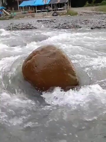 The video shows a view of a river with fast-moving water flowing over rocks. In the foreground, a large, round, brown rock is partially submerged in the water. In the background, there are trees and some structures that appear to be shops or stalls with blue and white awnings. Further in the distance, a metal bridge spans across the river. The sky is overcast.