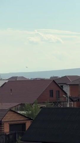 The video shows a view of several residential rooftops in the foreground. In the background, a distant landscape features rolling hills and a pale sky with some clouds. A small airplane is visible flying in the distance, over the hills. A brick tower-like structure is also visible on the left side of the background, behind some of the rooftops. The lighting suggests it is daytime.