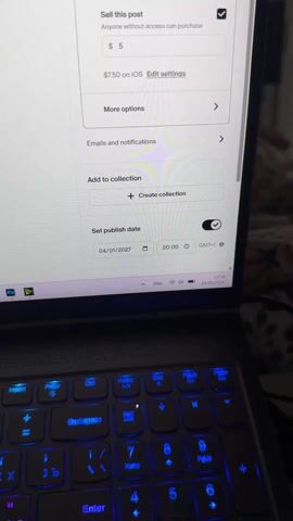 The video opens with a close-up shot of a laptop screen displaying a content creation platform, likely Patreon, showing options for a post. The text "Sell this post" is visible with a checkbox, and options to set a price of "$5" and "$7.50 on iOS" are shown. Below, there are sections for "More options," "Emails and notifications," and "Add to collection" (with a "Create collection" button). A prominent section is "Set publish date," which is toggled on. The date "04/01/2027" is set, along with the time "20:00 GMT+1." A finger with red nail polish points to the "04/01/2027" date. The Windows taskbar at the bottom of the screen indicates the current date as "24/03/2026." The laptop's keyboard is visible in the foreground, illuminated with green and pink backlighting. A female voice states, "I'm scheduling a post on my Patreon for 2027."

The camera then slowly pans to the right, revealing a bedroom interior. A white, soft, textured blanket is draped over a bed in the foreground, with a black cable lying on it. In the background, against a plain white wall, a small wooden table holds an electric space heater with its elements glowing a bright orange, suggesting it is active and producing heat. Beside the heater, a small, dark blue and white device, possibly a humidifier or air freshener, is placed. To the left of the heater, a window is partially visible, covered by a gray curtain adorned with artificial pink floral garlands. The overall lighting in the room appears dim, with the heater casting a warm glow.