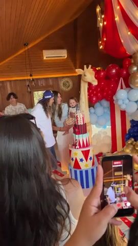 A birthday celebration is underway. The scene is decorated with a circus theme, evident in the red, white, and blue balloons and the multi-tiered cake adorned with circus imagery. A large giraffe decoration stands behind the cake. In the foreground, a person is holding up a smartphone, recording the event. A woman is holding a baby who is positioned next to the cake. To the left, a man is clapping enthusiastically, facing the woman and baby. Behind them, another young person is visible. The environment appears to be an indoor space with wooden walls and ceiling. The overall atmosphere is festive.