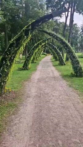 A dirt path winds through a park, lined on both sides by arched structures covered in dense green foliage. These arches create a tunnel effect over the path. Beyond the arches, trees and grass are visible, indicating an outdoor setting. The lighting suggests daytime. There are a few blurred figures walking on the path in the distance.