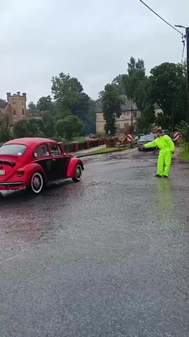 A person in a bright yellow jacket and pants stands on the right side of a wet asphalt road, facing a red Volkswagen Beetle. The car is driving away from the camera, turning slightly to the right. The person is gesturing with their right arm, pointing towards the direction the car is traveling. The road is lined with trees and buildings in the background. The sky is overcast, indicating a rainy or gloomy day. There is a wooden fence on the left side of the road.