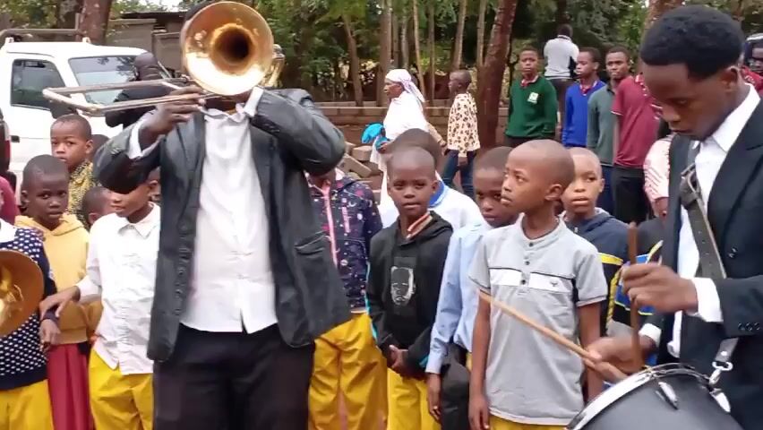 Here is a description of the video:

The video depicts three adult men, each playing a brass instrument (trumpets or trombones), positioned in front of a crowd of children. The men are dressed in formal attire, including white shirts and blazers, while the children are dressed in a mix of casual clothing, some wearing yellow pants. The performance is taking place outdoors in a grassy area, with vehicles visible in the background. The men appear to be engaging the children, who are watching attentively. The overall scene conveys a sense of musical performance and communal engagement.