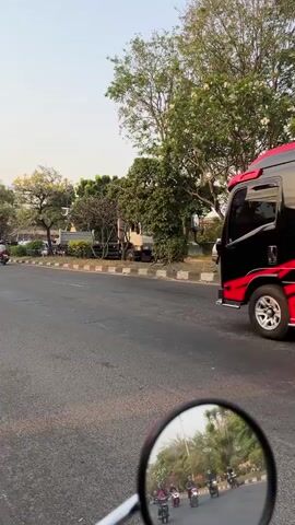 A black van with red and gold decals drives down a paved road. The van has images of a figure that appears to be Hanuman on its side. The road is lined with trees and bushes. There are several motorcycles on the road, some with riders and some with passengers. The scene is filmed from the perspective of someone on a motorcycle, with a partial view of the rider's mirror in the foreground. The sky is clear and the sun is shining.