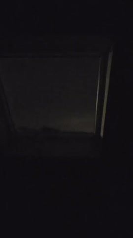 A video shows a dark interior with a window. The window displays a dark, cloudy sky. The clouds appear to be moving or shifting, with some light visible through them. The overall lighting in the video is very dim, suggesting it is nighttime or early morning. The camera is positioned low, looking upwards towards the window.