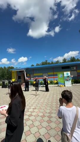 This media file captures an outdoor performance in Duvankul', Russia, under a bright, sunny sky with fluffy white clouds. The scene appears to be in front of a colorful building with a blue roof and yellow and blue striped walls, possibly a community center or an establishment associated with recreation given the banner on the right side.

The main subjects are a group of three performers dressed in black attire. Two women and one man are on a paved courtyard, performing for an unseen audience. The performers are actively engaged, with the women gesturing and singing, and the man holding a microphone and also seemingly singing or speaking. There is a music stand in front of the man.

In the foreground, a young woman with long dark hair is visible, holding a small white plate with some pink substance on it, possibly food or a craft material, and appears to be eating from it. Another individual in a white t-shirt is visible on the right, with their back to the camera, holding up a phone, suggesting they are recording or taking pictures of the event.

The overall atmosphere seems lively and festive, suitable for a daytime outdoor event. The weather is clearly pleasant and warm, indicated by the clear blue sky. The ground is paved with interlocking stones, adding to the organized but informal outdoor setting. The banner on the right side of the building has text in Russian, including "Урал" (Ural), which could indicate a regional connection or sponsor. The building itself seems to have windows and an entrance, suggesting it's an active facility.