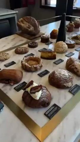 The video shows a display case filled with a variety of baked goods on a marble countertop with a gold-colored metal frame.  The display includes a large round loaf of bread, various pastries such as croissants, danishes, and tarts, and small cakes.  Each item has a small black label placed near it.  A tall, black, conical object sits among the pastries.  The background includes a grey counter, a fire extinguisher, and a window.  The camera slowly pans across the display, highlighting the different pastries.  A sign reads "Hands off, Please."