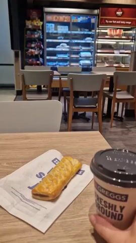 A hand holds a Greggs coffee cup in front of a table. On the table, next to the coffee cup, is a sausage roll on a Greggs napkin. In the background, a store interior with refrigerators stocked with food is visible.