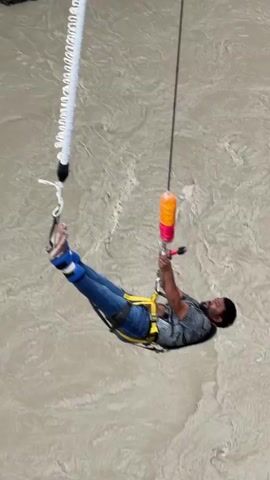 A person is bungee jumping over a river. The person is wearing blue jeans and a dark shirt. They are attached to a bungee cord that is connected to a platform above. The river is murky brown and appears to be flowing through a canyon. The canyon walls are rocky and steep. The sky is overcast.