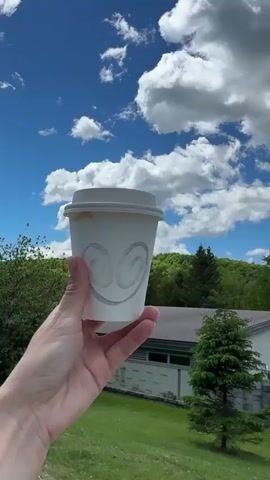A close-up shot shows a person's hand holding a white paper coffee cup with a lid. The cup features a subtle, embossed design resembling two swirling shapes. The backdrop is a vibrant blue sky dotted with fluffy white clouds. Below, there is a landscape of green trees, a patch of green grass, a parking lot with several parked cars, and a building. The person's hand is light-skinned and clad in a denim jacket. The video appears to be taken outdoors on a sunny day.