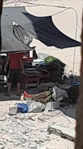 The video shows a person in a red shirt and dark shorts interacting with items under a makeshift shelter. The shelter appears to be constructed from tarps, one grey and one black, supported by unseen structures. Various items are scattered around the ground, including bags, what looks like a child's blanket or sleeping bag, and what could be clothing. In the background, there are stacks of what appear to be cardboard boxes or similar items. The ground is covered in gravel or small stones. The scene suggests an outdoor environment, possibly a campsite or a temporary dwelling.
