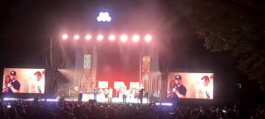 A wide shot of a live concert stage at night. A crowd of people is visible in the foreground, facing the stage. The stage is illuminated with bright pink and white lights. Several performers are on stage, some standing in a line, others scattered. There are two large screens on either side of the stage, displaying close-ups of individuals. Above the stage, a cluster of white lights forms a design. Banners hang from the stage structure. The overall atmosphere appears to be energetic and festive.