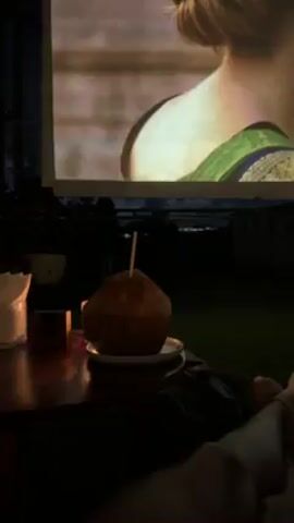 The video depicts an outdoor movie setting at night. The film playing on the screen features Julia Roberts in a scene where she is embracing a man in a white shirt. In the foreground, there is a wooden table with a young coconut drink, a small candle, and what appears to be a coaster. Two pairs of bare feet are also visible in the lower part of the frame. The background features a dark, indistinct view of buildings or vegetation. The overall atmosphere suggests a relaxed, intimate movie night experience.