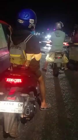 The video captures a nighttime scene of a traffic jam from the perspective of a motorcycle rider. The rider is wearing a blue helmet with stickers, a black hoodie with yellow accents, and shorts. A backpack with yellow accents is on their back. They are wearing sandals. The motorcycle has its taillight illuminated and a license plate visible. In front of the rider, another motorcycle is stopped, with a rider wearing a light green hoodie and a helmet, also carrying a backpack. Several other vehicles and motorcycles are visible in the background, with their lights on. The road appears wet, suggesting recent rain. The overall impression is of a slow-moving or stopped traffic situation at night, illuminated by vehicle lights and streetlights in the distance.