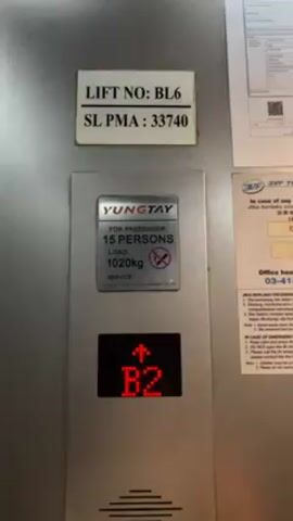 The video shows the control panel of an elevator in a building. The panel is made of brushed metal and has a display indicating the current floor as well as a set of stickers with information on the elevator. The top sticker reads "LIFT NO: BL6" and "SL PMA: 33740." Below that is a sign saying "YUNGTAY" and "FOR PASSENGER" with a "15 PERSONS" capacity, and "LOAD: 1020kg." Beneath that is the floor indicator which changes from "B2" to "B1" to "LG," along with an up arrow. A third sticker on the right side shows emergency information. The lighting in the elevator is bright and even. There are no people visible in the video, and the focus is entirely on the elevator panel.