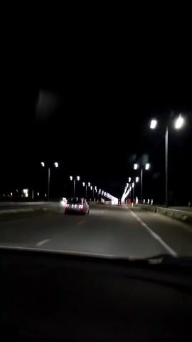 This video shows a first-person perspective of driving on a highway at night. The road is illuminated by numerous streetlights and the headlights of a few other vehicles. The asphalt road is divided into multiple lanes by white lines. The sky is dark, indicating nighttime. The perspective is from inside a vehicle, showing the dashboard or hood of the car in the foreground. Several cars are visible on the road ahead, including one red car in the left lane. The video is slightly shaky, suggesting movement.