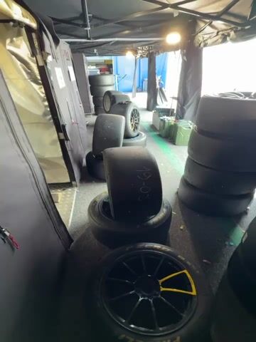 The video shows a storage area, possibly in a pit stop garage, filled with stacks of tires on wheels. The tires are black and appear new or lightly used. Some have numbers written on them in white marker, like "19-03".  The wheels are mostly black with some yellow accents on the spokes.  There are several stacks of tires, some leaning against walls or covers, others standing upright. The lighting is bright but uneven, and the area is cluttered. A metal hand truck is visible behind some tire stacks.  The space is enclosed by dark tents or coverings.